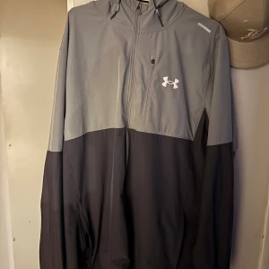 Grå och svart vindjacka Under Armour XL - Snygg tvåfärgad vindjacka från Under Armour i grått och svart. Jackan har huva, halv dragkedja framtill, bröstficka med dragkedja och Under Armour-logga på bröstet. Perfekt för dig som gillar sportig stil och vill ha något lätt och funktionellt.