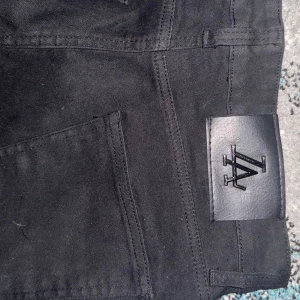 Svarta jeansbyxor från Karl Kani - Svarta jeansbyxor från Karl Kani med klassisk femficksdesign och svart läderpatch med logga bak i midjan. Byxorna har raka ben och är tillverkade i ett slitstarkt jeanstyg. Perfekta för dig som gillar streetwear och enkel stil.