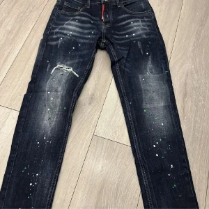 Mörkblå Dsquared2 jeans  - Snygga mörkblå jeans från Dsquared2 med coola färgstänk i vitt och grönt, slitningar och broderad detalj bak. Klassisk femficksmodell med raka ben och kontrastsömmar. Perfekta för dig som vill sticka ut med en unik streetstyle.