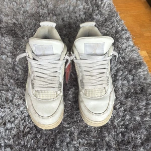 Nike Air Jordan 4 Retro Vit - Säljer ett par Nike Air Jordan 4 Retro i helvitt skinn med silvriga detaljer och meshpaneler. Klassisk siluett med snörning, synlig Air-enhet i sulan och Jumpman-logga på hälen. Perfekta för dig som gillar streetwear och sneakers med ikonisk design.