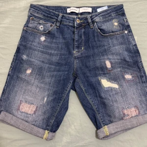 Blå jeansshorts från Gabba - Snygga blå jeansshorts från Gabba med slitna detaljer och lagningar för en cool, avslappnad vibe. Klassisk femficksmodell, uppvikta benslut och kontrastsömmar. Perfekt för dig som gillar streetstyle och vill ha något som sticker ut.