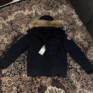 Canada Goose wyndham - Säljer en mörkblå Canada Goose dunjacka med huva och snygg fuskpälsdetalj. Jackan har klassisk patch på ärmen, flera fickor och stängs med både dragkedja och knappar. Perfekt för kalla vinterdagar och riktigt stilren look.