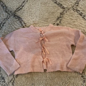 Rosa kofta med rosetter och knyt - Supergullig ljusrosa kofta med ribbad struktur och fyra söta rosetter framtill som knäppning. Långärmad och croppad modell, perfekt för lager på lager. Mjuk och mysig känsla, passar till både jeans och kjol.