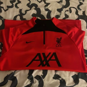 Liverpool Nike träningsöverdel röd - Röd långärmad träningsöverdel från Nike med Liverpool FC-logga och AXA-tryck på bröstet. Svarta detaljer vid axlar och dragkedja framtill. Perfekt för fotbollsträning eller som supporterplagg. Denna tröja sitter ganska så tajt då det är en träning tröja. Så om du har storleken L eller M passar den dig galant. Priset kan diskuteras.