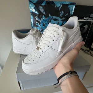 Nike Air Force 1 vita sneakers - Klassiska Nike Air Force 1 sneakers i helvitt skinn med perforerad tå, rund tå och platt sula. Snygg och tidlös modell med snörning och diskret Nike-logga på sidan. Perfekta för dig som vill ha en clean och stilren look till vardags.