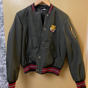 Mörk Grön bomberjacka med tigerpatch - Snygg mörkgrön bomberjacka från H&M med broderad tigerpatch på bröstet. Jackan har röda och svarta ränder på muddar och krage, dragkedja framtill och fickor med lock. Ärmarna är långa och jackan har en klassisk bomberpassform.