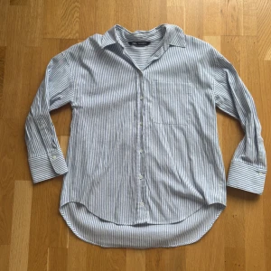 Snygg randig skjorta från Zara - Snygg skjorta från Zara i vitt och blått med tunna vertikala ränder. Klassisk krage, bröstficka och knäppning framtill. Skjortan är tillverkad i en mjuk blandning av  och polyester, perfekt för en chill och stilren look. I väldigt fint skick, knappt använd. 