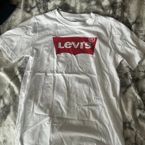 Vit Levi's t-shirt med röd logga - Vit t-shirt från Levi's med klassisk röd logga tryckt på bröstet. T-shirten har rund halsringning och korta ärmar. Tillverkad i mjuk bomull för en skön känsla och enkel stil den är helt ny lite vikt för den har ståt i paketet men annars är den jätte fin och bra skick mvh Omar 