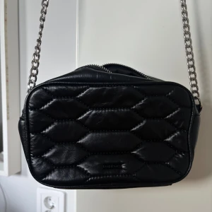 Svart quiltad axelväska från Parfois - Snygg svart axelväska från Parfois i quiltat skinnimitation. Väskan har silverfärgad kedja och justerbart axelband i läderlook. Perfekt storlek för mobil och småsaker, med dragkedja upptill och stilren design.
