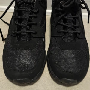 Svarta sneakers med snörning - Svarta sneakers från Freaks med klassisk design och snörning. Ovandelen är i en mix av syntet och mocka, med detaljer i svart och diskret logga på sidan. Skorna har platt sula med rutmönster undertill och rund tå. Perfekta för en stilren och sportig look.