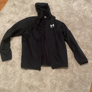 Svart vindjacka från Under Armour M - Svart vindjacka från Under Armour med huva och dragsko. Jackan har dragkedja framtill och meshfoder på insidan för extra ventilation. Perfekt för träning eller när du vill ha en sportig look. Under Armour-logga i vitt på bröstet. Passar även S. Endast swish skickas spårbart 59kr