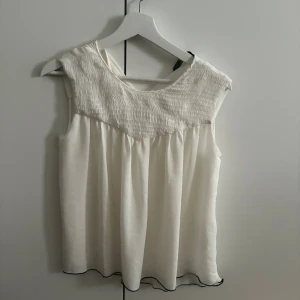 Vit kortärmlös blus - Säljer en luftig vit blus från Zara med smockad överdel och lätt rynkad nederdel. Blusen är ärmlös och har en flowy passform med diskret svart kant längst ner. Perfekt för varma dagar och enkel att styla till olika looks.