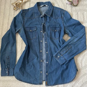 Blå jeansskjorta från Lindex - Snygg blå jeansskjorta från Lindex med klassisk krage, tryckknappar och två bröstfickor med lock. Skjortan har lång ärm och markerade sömmar för en cool denimlook. Perfekt att styla lager på lager eller som den är.