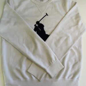 Vit sweatshirt från Polo Ralph Lauren - Vit sweatshirt från Polo Ralph Lauren med stor svart broderad polospelare på bröstet. Klassisk rund halsringning och långa ärmar. Tillverkad i mjuk bomull för en skön och avslappnad stil.