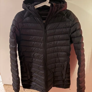 Svart dunjacka Peak Performance S - Svart jacka från Peak Performance i storlek S. Jackan har huva, dragkedja framtill och två sidofickor. Snyggt quiltad design med diskret broderad logga på bröstet. Perfekt för kyliga dagar och enkel att matcha med allt.Ett litet hål vid ärmen som är enkel att sy eller lägga en patch på.