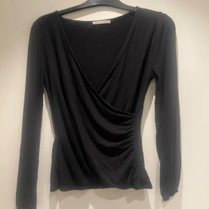 Svart omlottopp från Anna Field - Svart långärmad omlottopp från Anna Field med v-ringning och lätt rynkade detaljer i sidan. Toppen har smala muddar vid ärmsluten och är tillverkad i mjuk bomullsmix. Perfekt för dig som gillar en stilren och enkel look.