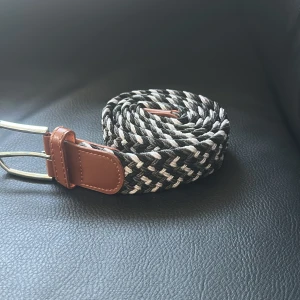 Svartvitt flätat bälte med skinn-detaljer - Snyggt flätat bälte i svart och vitt med bruna skinn-detaljer vid spännet och änden. Bältet har ett klassiskt silverfärgat spänne och är gjort i en mix av textil och skinn. Perfekt för att ge outfiten en cool touch.