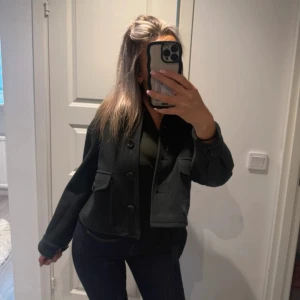 Svart croppad overshirt med fickor - Snygg svart croppad overshirt med stora knappar och två rejäla fickor framtill. Jackan har en loose passform och är långärmad, perfekt för lager-på-lager. Materialet känns mjukt och ger en clean look.