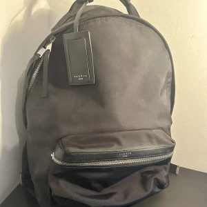 Svart ryggsäck från Sandro Paris - Stilren svart ryggsäck från Sandro Paris med praktiskt ytterfack med dragkedja💫 Perfekt för dig som vill ha en snygg och funktionell väska till skolan eller jobbet🍁Väskan är sparsamt använd, i väldigt fint skick. Nypris ligger på runt 3000 kr. Mitt pris är 999 kr utgångspris