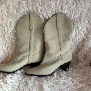 Isabel Marant ljusa cowboy boots - Snygga cowboy boots från Isabel Marant i ljusbeige mocka och skinn. Boots med spetsig tå, markerad klack och klassiska western-detaljer. Kommer med originalkartong, dustbag och extra klackar. Perfekta för dig som vill sticka ut med stil.