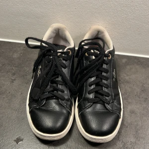 Svarta sneakers från Björn Borg - Svarta sneakers från Björn Borg med vita sulor och svarta snören. Skorna har detaljer i guld på hälen och logga på sidan. Ovandelen är i skinn och insidan är fodrad i vitt tyg. Klassisk rund tå och platt sula för en clean look. Använd några ggr och slitna därför ett billigt pris
