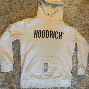 Vit hoodie från Hoodrich med tryck - Vit hoodie från Hoodrich med stor svart logga framtill och coola tryck på ryggen och ärmen. Klassisk känguruficka och huva med snörning. Mjuk bomullskänsla och streetwear-vibe, perfekt för dig som gillar stilrena men ändå unika plagg.