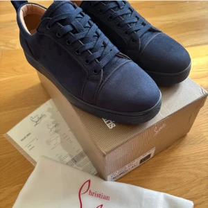 Mörkblå sneakers från Christian Louboutin - Snygga mörkblå sneakers från Christian Louboutin i mocka med klassisk röd sula och rund tå. Skorna har snörning och svart platt sula, perfekt för dig som vill sticka ut med exklusiva detaljer och stilren design.