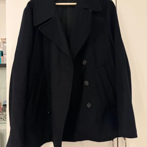 Svart skepparkavaj med knappar - Klassisk svart skepparkavaj med dubbelknäppning och bred krage. Jackan har två snedställda fickor framtill och är tillverkad i ett tjockare material som passar perfekt för kyligare dagar. Enkel och stilren design som funkar till det mesta.