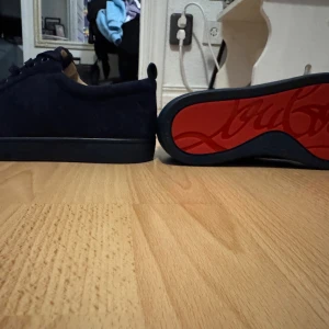 Christian Louboutin marin blå sneakers - Säljer ett par svarta sneakers från Christian Louboutin i mocka med den ikoniska röda sulan. Skorna har snörning och en stilren, låg profil. Kommer med originalkartong, dustbag och tillbehör. Perfekta för dig som vill sticka ut med lyxig känsla. Allt på bilden ingår, helt nytt skick aldrig använt.