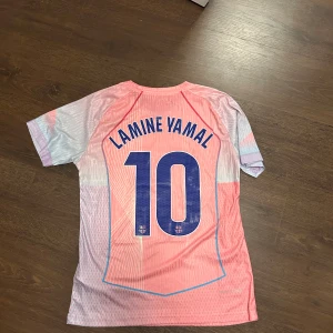 Barça Lamine Yamal rosa matchtröja - Säljer en FC Barcelona fotbollströja med Lamine Yamal och nummer 10 på ryggen. Tröjan är rosa med ljusblå och vita detaljer, Nike-logga och klubbmärke framtill samt Spotify som sponsor. Tillverkad i lätt och ventilerande material, perfekt för match eller träning. Helt ny