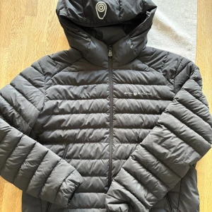 Svart pufferjacka från Sail Racing - Säljer en svart pufferjacka från Sail Racing med huva och dragkedja framtill. Jackan har horisontella sömmar, två sidofickor och logga på bröstet och huvan. Perfekt för kalla dagar och riktigt snygg streetstil.