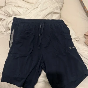 Marinblå shorts från BOSS - Snygga marinblå shorts från BOSS med diskret logga på benet och snörning i midjan. Shortsen har elastisk midja och grå detalj vid fickorna. Perfekta för en avslappnad och sportig look.
