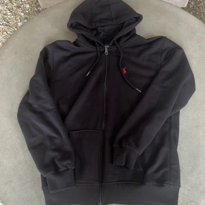 Svart hoodie från Polo Ralph Lauren  - ⚠️OBS står fel på etiketten, storleken är M‼️Svart hoodie från Polo Ralph Lauren i storlek XL. Klassisk modell med dragkedja, huva med snörning och känguruficka. Broderad röd logga på bröstet. Mjuk insida och ribbade muddar. Perfekt för chill dagar eller när du vill ha en clean streetstyle.