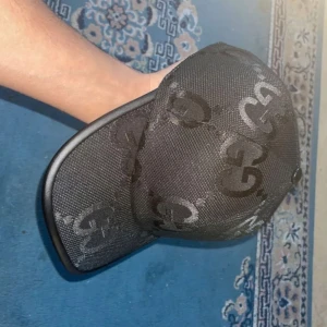 Svart keps från Gucci med GG-mönster - Säljer en svart keps från Gucci med klassiskt GG-mönster i mörkgrått. Kepsen har böjd skärm och justerbar rem bak. Tillverkad i slitstarkt material med en lyxig känsla, perfekt för dig som vill ha en stilren accessoar med designer-vibe.