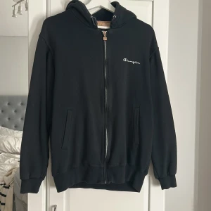 Svart hoodie från Champion med dragkedja - Svart hoodie från Champion i storlek M. Klassisk modell med huva, hel dragkedja framtill och två fickor. Mjuk och skön, perfekt för chill dagar. Diskret Champion-logga broderad på bröstet. Passar dig som gillar enkel och stilren streetwear.