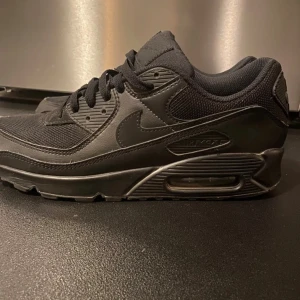 Nike Air Max 90 - Säljer ett par helsvarta Nike Air Max 90 sneakers med klassisk siluett. Skorna har en mix av skinn och mesh, synlig Air Max-dämpning i sulan och snygga detaljer i svart. Perfekta för dig som gillar en clean och sportig look.