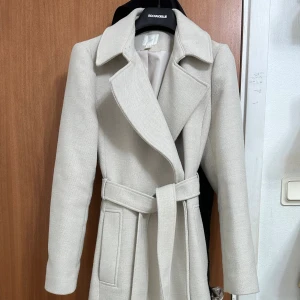 Beige kappa med bälte från H&M - Snygg beige kappa från H&M med knytskärp i midjan. Lång modell med raka ärmar och diskreta fickor på sidorna. Perfekt för dig som vill ha en stilren och klassisk look under höst och vinter. 