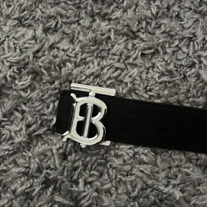 Burberry bälte med TB-spänne - Säljer ett Burberry bälte med ikoniskt TB-spänne i silverfärgad metall. Ena sidan är svart med diskret rutmönster och präglad logga, andra sidan är beige med klassiskt Burberry-rutmönster i rött och svart. Bältet är tillverkat i skinn och har en stilren, lyxig vibe.