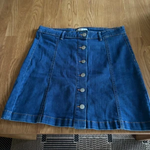 Blå jeanskjol med knappar från g perfect jeans - Snygg blå jeanskjol från g perfect jeans i storlek 40. Kjolen är kort och har en A-linjeform med fem silverfärgade knappar framtill och hällor för bälte. Tillverkad i klassisk denim med kontrastsömmar som ger en cool vibe.