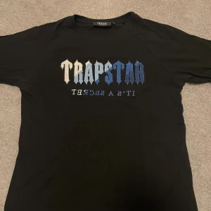 Trapstar svart t-shirt med blått tryck - Svart t-shirt från Trapstar i storlek L med stort broderat Trapstar-tryck i gradient från vitt till blått på bröstet. Rund hals och klassisk passform. Materialet är mjuk bomull och trycket har en cool textstil som sticker ut.