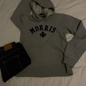 Grå hoodie från Morris Vintage - Snygg grå hoodie från Morris med svart tryck framtill och klassisk känguruficka. Tröjan har huva med dragsko och mjukt innerfoder. Perfekt för chill dagar och enkel att matcha med jeans eller joggers.