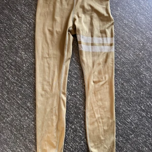 Beige leggings från Stronger med ränder - Snygga beige leggings från Stronger med två vita randiga detaljer på ena benet. Tillverkade i stretchigt material av 75% återvunnen polyester och 25% elastan. Perfekta för träning eller chill dagar, med tight passform och bekväm midja.