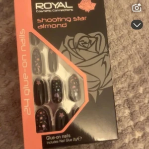 Royal Shooting Star Almond lösnaglar - 24 svarta glue-on lösnaglar i almond-form med stjärnor, månar och prickar i lila, rosa och blått. Paketet innehåller nagellim (2g). Perfekt för dig som vill ha en unik och färgglad nageldesign med cosmic vibes. Helt oanvända