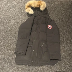Svart dunjacka Canada Goose med päls - Säljer en svart dunjacka från Canada Goose med ikonisk patch på ärmen och stor huva med beige päls. Jackan har flera fickor med lock, tryckknappar och dragkedja. Perfekt för kalla vinterdagar och riktigt snygg streetstil.