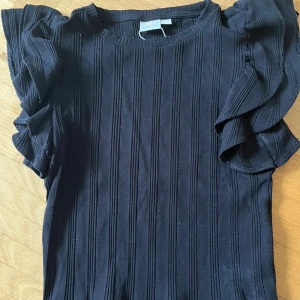 Svart ribbad topp med volangärm - Snygg svart ribbad topp från Gina Tricot med volangdetaljer på ärmarna. Toppen har rund halsringning och är tillverkad i ett stretchigt material som sitter skönt på kroppen. 