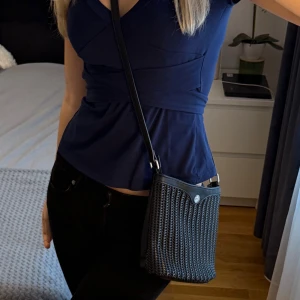 Svart axelväska från Don Donna - Snygg svart axelväska från Duffy med flätad framsida och detaljer i skinn. Väskan har ett silverfärgat emblem framtill, justerbar axelrem och dragkedja på baksidan. Perfekt storlek för mobil och småsaker, stilren och enkel att matcha. Har en liten missfärgning på underdelen av väskan (se bild 4).