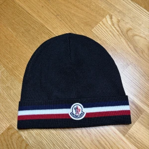 Svart Moncler mössa - Svart stickad mössa från Moncler med klassisk Moncler-logga framtill. Upptill är mössan enkel och stilren, medan kanten har snygga ränder i rött, vitt och marinblått. Tillverkad i 100% ull för extra värme och komfort. Perfekt för dig som gillar exklusiva accessoarer.