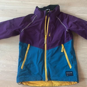 Färgglad vindjacka Polarn O. Pyret 116 - Säljer en snygg vindjacka från Polarn O. Pyret i storlek 116. Jackan har lila och blå färgblock, gula dragkedjor och reflexdetaljer. Perfekt för blåsiga dagar och har meshfoder på insidan. Jackan har hög krage och två fickor framtill.