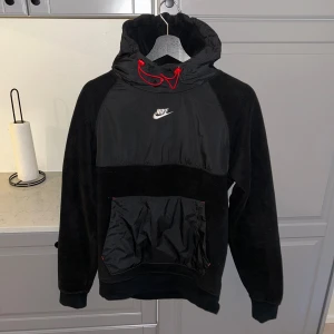 Svart hoodie från Nike med röd snörning - Svart hoodie från Nike med fleece och syntetmaterial, stor magficka och huva med röd snörning. Nike-logga broderad på bröstet. Perfekt för dig som gillar sportig och chill stil. (passar s)