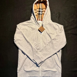 Grå hoodie från Burberry med dragkedja - Snygg grå hoodie från Burberry med klassiskt rutigt foder i huvan. Tröjan har dragkedja framtill, snörning vid huvan och subtilt Burberry-tryck på bröstet. Tillverkad i mjuk bomull, perfekt för chill dagar. Långärmad och med praktiska fickor på sidorna.